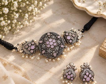May include: A silver-toned choker necklace featuring a large circular centerpiece and two side panels, all inlaid with pale pink stones and accented with small white beads. The necklace includes a black cord and matching stud earrings.