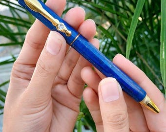 May include: A ballpoint pen with a deep blue barrel featuring gold-tone metal accents. The pen includes a gold clip with a small clear stone, a gold-tone textured top button, and a gold-tone conical tip. The barrel has a smooth, polished finish.