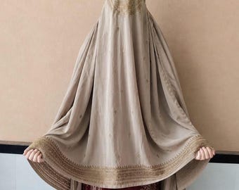 May include: A beige burqa with intricate gold embroidery around the face covering and hem. The garment drapes to the floor, revealing a glimpse of a maroon and white patterned garment underneath. The burqa is a traditional garment.