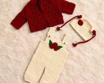 May include: A handmade baby outfit set, including a red knit jacket with bobble details, cream-colored overalls with red flower accents, and a matching hat with tassels. The set is displayed on a soft, white surface.