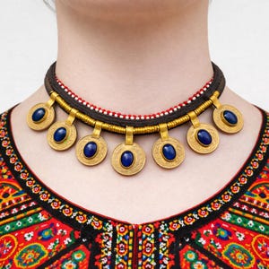 May include: A gold necklace with seven circular pendants, each with a blue oval stone. The necklace has a dark brown band with red and white beaded accents. The person is wearing a colourful embroidered top.