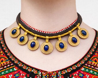 May include: A gold-toned necklace with seven circular pendants, each featuring a blue oval stone. The necklace has a dark brown band with red and white beaded accents. The person is wearing a colorful embroidered top.