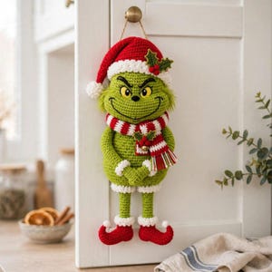 Könnte beinhalten: Eine gehäkelte grüne Charakterpuppe mit einer rot-weißen Weihnachtsmannmütze mit Bommel, einem passenden rot-weiß gestreiften Schal mit Stechpalmen-Akzent und roten Schuhen mit aufgebogenen Spitzen und weißen Manschetten.