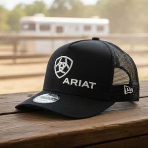 May include: Black baseball cap with a white Ariat logo and text. The cap has a mesh back and a curved brim. The New Era logo is on the side. The cap is sitting on a wooden surface.