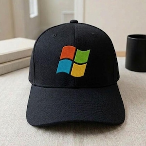 May include: Black baseball cap featuring the Windows logo in a colorful design. The logo includes a red, blue, green, and yellow square. The cap is on a light-colored surface, with a black mug and books in the background.