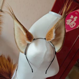 May include: A thin black metal headband featuring two large, pointed faux fur animal ears. The ears are colored in shades of brown, tan, and cream, with long, wispy tufts of hair extending from the tips. The ears are positioned upright on the band.