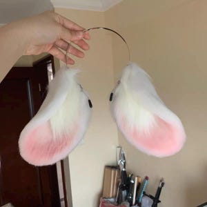 Puede incluir: Un par de orejas de animal de piel sintética blanca y rosa claro unidas a una diadema de metal fina y flexible. Las orejas presentan un pelaje blanco largo y suave en los bordes exteriores y un pelaje rosa claro denso en el interior.