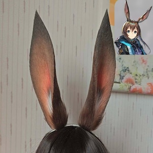Op de afbeelding: Een paar hoge, puntige, donkerbruine dierenoren met rood-oranje binnenschaduw en witte vachtaccenten aan de basis. De oren zijn bevestigd aan een donkere haarkleurige basis, en een kleine illustratie van een anime-figuur is zichtbaar.