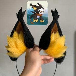 May include: A pair of pointed animal-style ears attached to a thin headband. The ears feature black outer edges, brown inner sections, and bright yellow faux fur tufts at the base. Above the ears is a small square image of a cartoon creature.