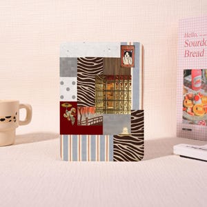 Könnte beinhalten: Ein rechteckiger Notizbuchumschlag mit einer Collage aus Mustern, darunter Zebrastreifen, vertikale blau-weiße Streifen, graue Punkte und ein Gitter aus goldenen Quadraten. Er enthält kleine Illustrationen einer Katze und einer Figur.