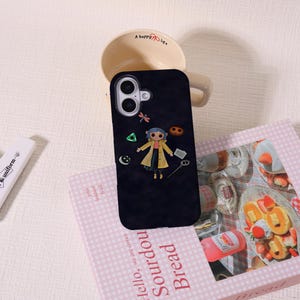 May include: Black phone case featuring a cartoon character in a yellow coat, with a dragonfly and other small icons. The case is resting on a pink book with the words "Hello, Sourdough Bread" and a coffee mug.