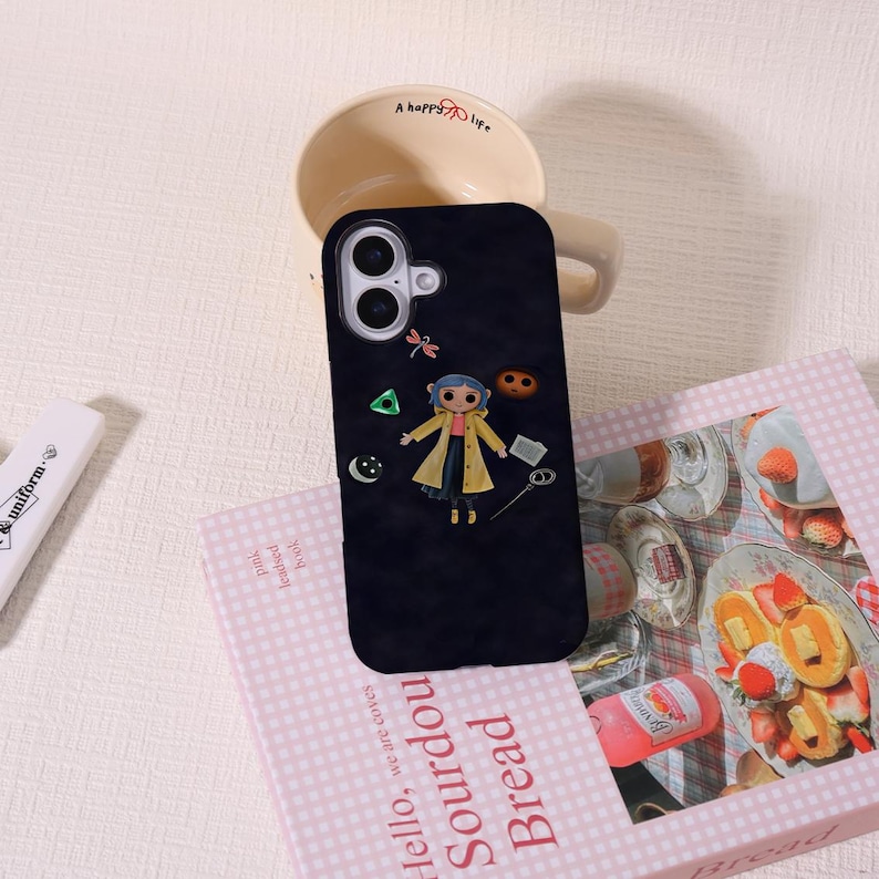 May include: Black phone case featuring a cartoon character in a yellow coat, with a dragonfly and other small icons. The case is resting on a pink book with the words "Hello, Sourdough Bread" and a coffee mug.