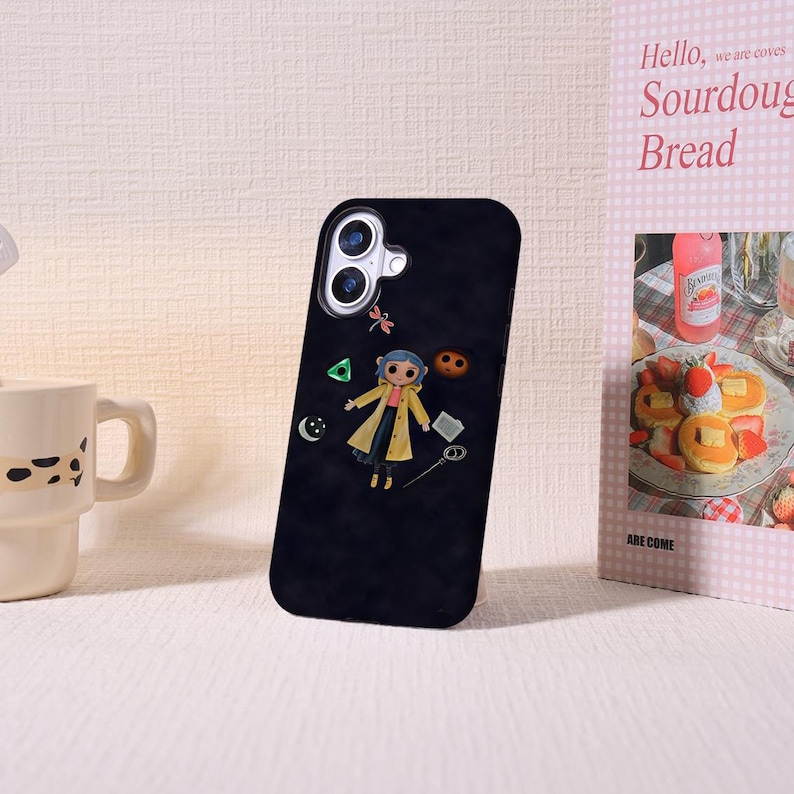 May include: Black phone case featuring a cartoon character in a yellow coat, with a dragonfly, a green triangle, and other small icons. The phone case is next to a mug and a pink and white book with the words "Hello, we are coves Sourdough Bread."