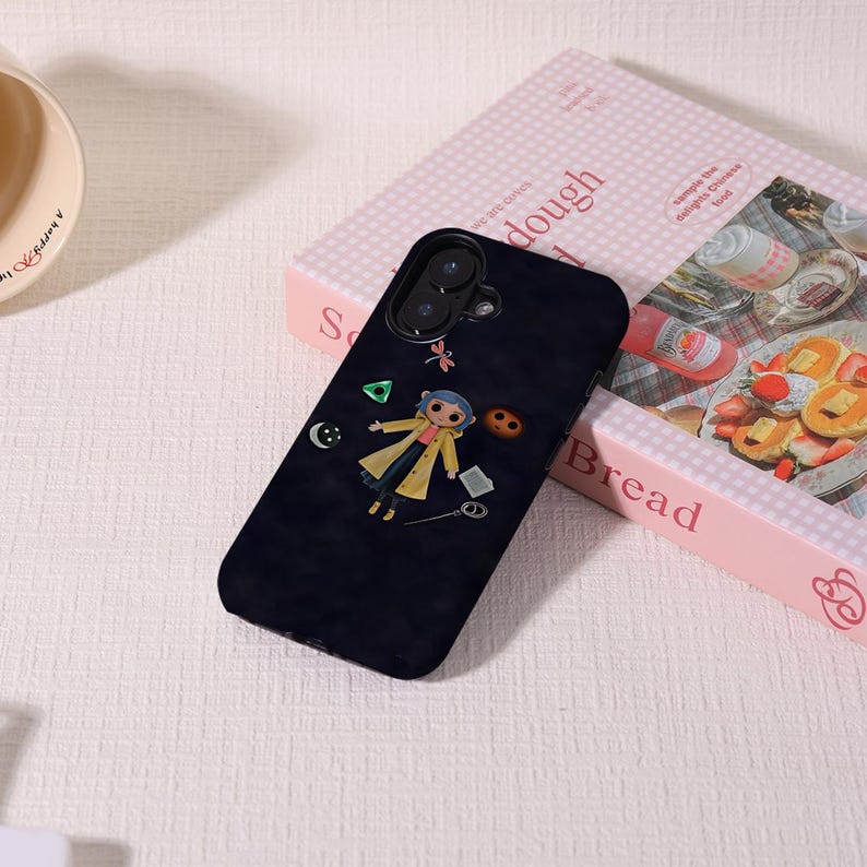 May include: A black phone case featuring a cartoon character in a yellow coat, with a blue hair and a variety of small, floating objects. The case is resting on a pink box with the word "dough" printed on it.