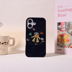May include: Black phone case featuring a cartoon character in a yellow coat. The case has a camera cutout and decorative elements including a dragonfly, a green triangle, and a crescent moon. A mug and a pink sign with the words "Sourdough Bread" are in the background.