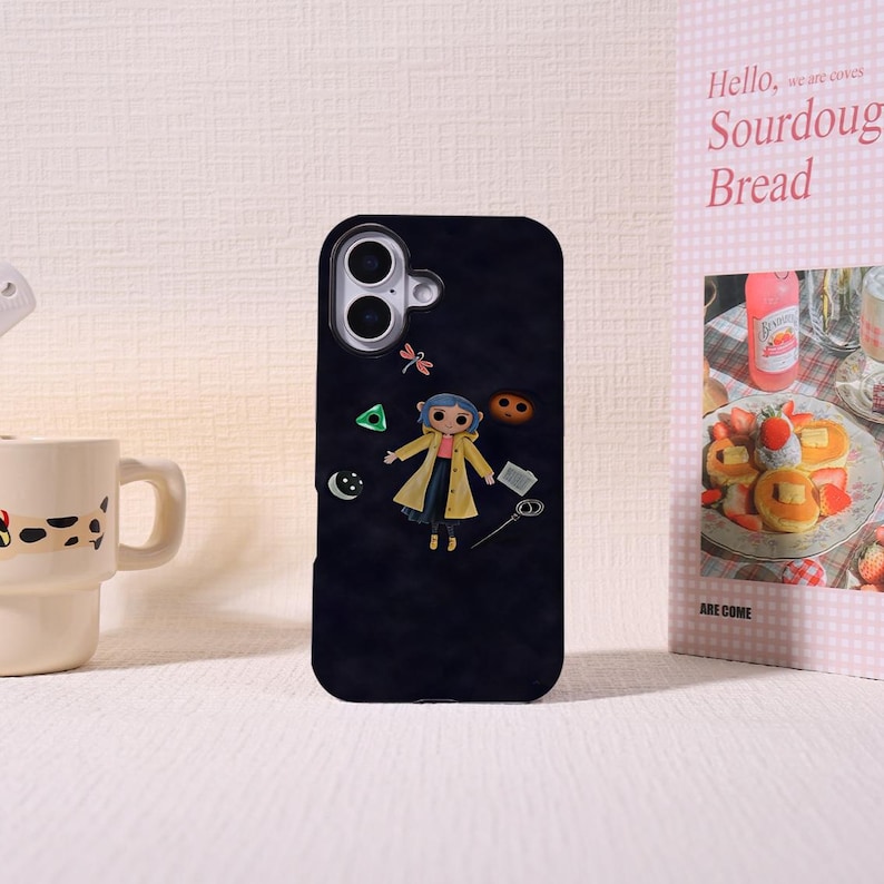 May include: Black phone case featuring a cartoon character in a yellow coat. The case has a camera cutout and decorative elements including a dragonfly, a green triangle, and a crescent moon. A mug and a pink sign with the words "Sourdough Bread" are in the background.