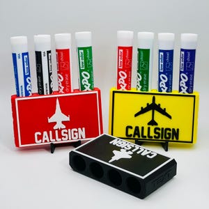 Könnte beinhalten: Ein Set aus drei Whiteboard-Markerhaltern in Rot, Gelb und Schwarz, jeweils mit einer Flugzeuggrafik und dem Wort "CALLSIGN". Die Halter sind für Whiteboard-Marker in verschiedenen Farben konzipiert.