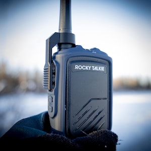May include: A dark blue Rocky Talkie walkie-talkie held in a gloved hand against a blurred winter landscape. The device has an antenna, a speaker, and the brand name printed on the front.