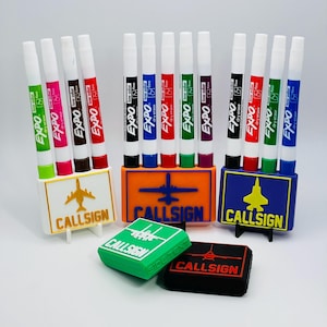 May include: A collection of Expo dry erase markers in various colors, displayed in custom holders with airplane and jet designs. The holders and erasers feature the text "CALLSIGN".