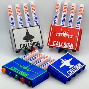 May include: A collection of whiteboard marker holders in various colors, each with a different aircraft silhouette and the word "CALLSIGN". The holders are paired with white Artline 500 whiteboard markers.