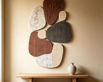 May include: A wall-mounted art installation composed of multiple organic, pebble-shaped panels in varying textures and colours, including dark brown, charcoal grey, cream, and marbled white, positioned above a simple light-toned wooden bench.