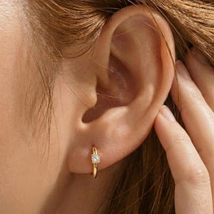Könnte beinhalten: Nahaufnahme eines goldenen Creolen-Ohrrings mit einem einzelnen, klaren Edelstein. Der Ohrring wird an einem Ohr getragen, wobei die Hand der Person ihre Haare berührt. Schmuck von Lunora Jewelry.