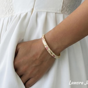 Peut inclure: Un bracelet jonc doré doté d'une surface en émail crème ornée de petits motifs floraux roses et de délicats motifs de volutes dorées. Le bracelet est porté au poignet, avec le texte Lunora Jewelry visible dans le coin de l'image.