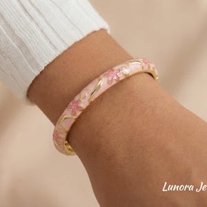 Puede incluir: Una pulsera brazalete de esmalte rosa y dorado con un diseño floral. La pulsera está adornada con flores rosas y detalles dorados. La joya se lleva en la muñeca y la marca "Lunora Jewelry" es visible.