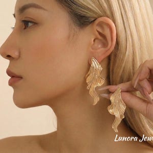 Puede incluir: Pendientes dorados texturizados con un diseño fluido y orgánico. Los pendientes se sostienen cerca de la oreja, mostrando su forma y detalle. Tienen una apariencia ondulada y en capas.