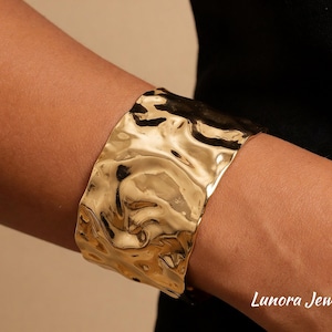 Può includere: Un bracciale a polsino largo, color oro, con finitura martellata e strutturata. Il gioiello è indossato su un braccio, con il logo Lunora Jewelry visibile in basso a destra.