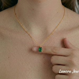 Puede incluir: Un collar dorado con un colgante rectangular verde esmeralda. El colgante está sujeto por un dedo. El collar está sobre una persona. El texto "Lunora Jewelry" está en la esquina inferior derecha.