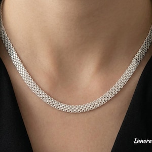 Könnte beinhalten: Eine silberfarbene Halskette mit einem gewebten Kettendesign. Die Halskette wird vor einem neutralen Hintergrund präsentiert, der ihre komplizierte Textur und reflektierende Oberfläche hervorhebt. Die Halskette ist im Choker-Stil.