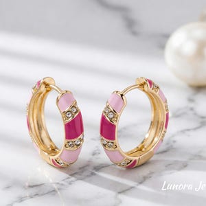 Puede incluir: Un par de pendientes de aro dorados con rayas alternas de esmalte rosa claro y oscuro. Los pendientes están adornados con pequeños cristales transparentes. Los pendientes se exhiben sobre una superficie de mármol.