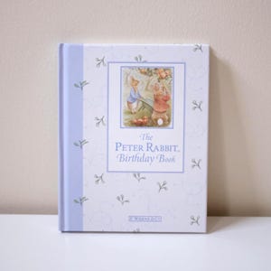 Peut inclure: Un livre relié intitulé The Peter Rabbit Birthday Book avec un dos violet clair et une couverture blanche ornée d'un motif répétitif de petits brins verts et d'une illustration centrale de lapins, avec l'éditeur F. Warne & Co. en bas.