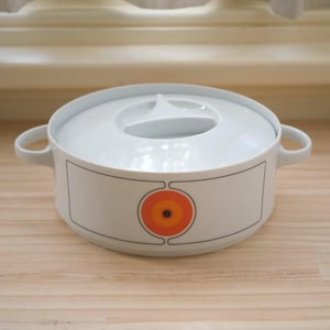 Puede incluir: Cazuela blanca con tapa y dos asas. El plato presenta un diseño rectangular con un gráfico circular naranja y amarillo. Está sobre una superficie de madera.