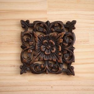 Może przedstawiać: Ciemnobrązowa, kwadratowa drewniana dekoracja ścienna z kwiatowym i zwijanym wzorem. Rzeźbiony element przedstawia centralny kwiat otoczony ozdobnymi, wirującymi wzorami. Widoczne usłojenie drewna dodaje tekstury elementowi dekoracyjnemu.