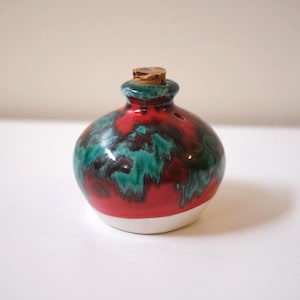 May include: A small, round ceramic bottle with a cork stopper. The bottle is painted with a vibrant red base, overlaid with teal and dark green patterns. The bottom is white.