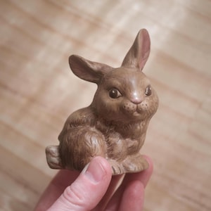 May include: A brown ceramic bunny figurine with detailed features, including upright ears with pink interiors, dark eyes, and a textured fur appearance. The bunny is in a seated position, showcasing its paws and overall form.