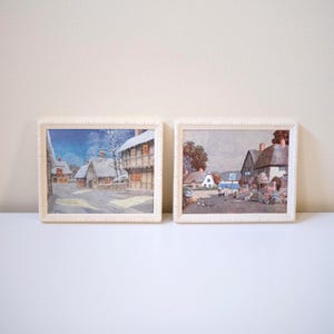 May include: Two rectangular framed prints featuring village scenes. The left print depicts a snow-covered street with timber-framed houses under a blue sky. The right print shows a village street with thatched-roof cottages and greenery.