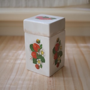 May include: A white ceramic square container with a lid, decorated with red strawberries, green leaves, and white flowers. The container is on a light-coloured wooden surface.