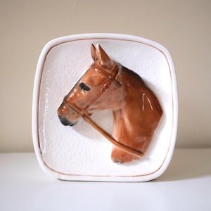 May include: A square ceramic wall plaque featuring a three-dimensional relief of a brown horse head in profile. The horse wears a brown bridle, and the background is a textured white surface with a thin brown border along the rounded edges.