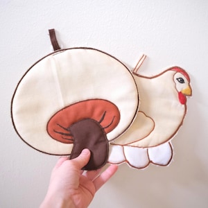 May include: A fabric pot holder shaped like a mushroom with a cream cap and brown stem, layered over a second fabric pot holder shaped like a white chicken with a red comb and yellow beak, both featuring brown trim and small fabric hanging loops.