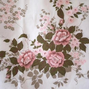 May include: A white fabric featuring a floral pattern with large pink roses and smaller pink blossoms. The design includes dark olive green leaves and stems, with a vertical vine pattern running along the right side of the textile surface.