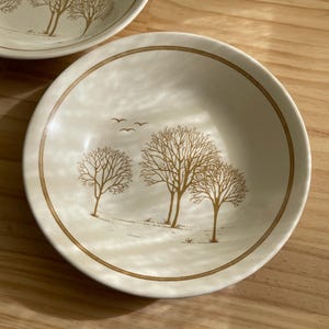 Puede incluir: Cuencos de cerámica blanquecina con un diseño de árboles y pájaros marrones. Los cuencos tienen un borde marrón y una escena de paisaje con árboles y pájaros. Los cuencos están sobre una superficie de madera.