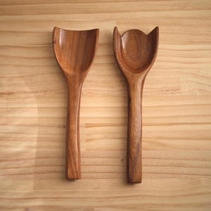 May include: Two wooden salad spoons with unique, curved designs. One spoon has a rounded, scoop-like head, while the other features a tulip-shaped head. Both are crafted from a warm, medium-toned wood and rest on a light-colored wooden surface.