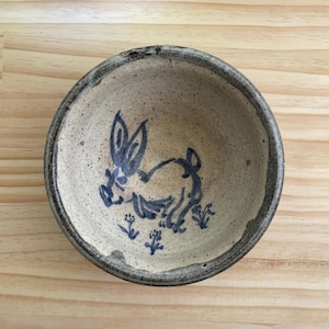 Könnte beinhalten: Eine kleine, runde Keramikschale mit einem handgemalten blauen Hasenmuster. Die Schale hat einen gesprenkelten beigefarbenen Innenraum und einen dunkelbraunen Rand. Der Hase ist in einem einfachen, skurrilen Stil mit floralen Akzenten dargestellt.