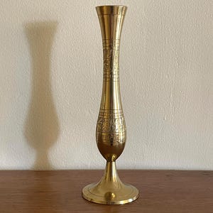 May include: A tall brass vase with a flared top and base. The vase has etched floral patterns around the middle section. The vase stands on a round base and is displayed on a wooden surface.