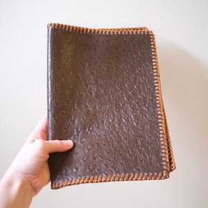 Puede incluir: Una carpeta rectangular de color marrón oscuro con una superficie texturizada que recuerda a la piel de avestruz. Los bordes están rematados con un detalle de puntada de látigo en color tostado claro que recorre el perímetro de la cubierta.
