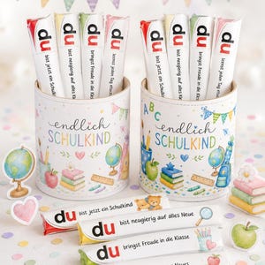 May include: Two cylindrical containers decorated with school-themed illustrations and the text "endlich SCHULKIND". Each holds four white candy bars labeled "du" followed by German phrases. Four additional candy bars lie horizontally in front.