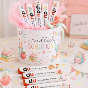 May include: A cylindrical gift box decorated with school-themed illustrations and the text "endlich SCHULKIND," filled with pink tulle and five chocolate bars labeled "du" with various phrases and icons, plus four additional chocolate bars in front.